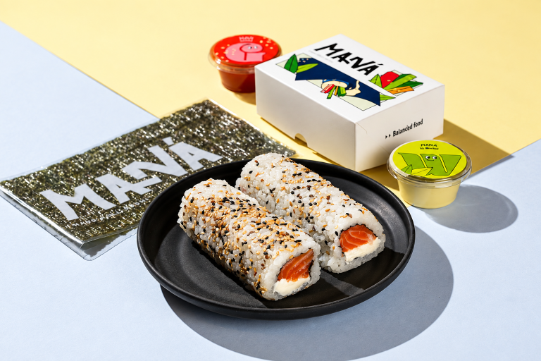 Hand rolls de salmão com cream cheese e gergelim em prato preto, acompanhados de nori e embalagens do Maná Hand Roll.